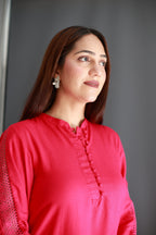 A woman with long brown hair wearing a bright pink long-sleeve top with textured side panels and a buttoned neckline, photographed against a dark gray background.