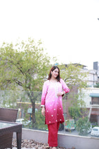 A woman stands on a rooftop terrace wearing a pink and red gradient traditional outfit with white floral patterns. The background features green trees, glass railings, and outdoor seating.