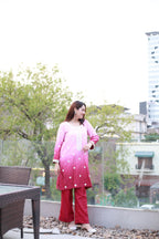 A woman stands on a rooftop terrace wearing a pink and red gradient kurta with white embroidered details and matching red pants. The background features green trees, a glass railing, and a city building under a bright, overexposed sky.