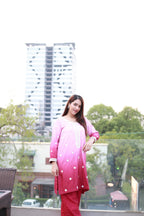 A woman modeling a pink and red gradient salwar kameez suit with white embroidered details, standing on a balcony with a modern building and trees in the background.