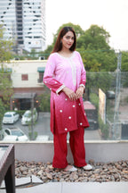 A woman modeling a pink and red two-tone kurta set with white embroidery, standing on a rooftop with a city background.