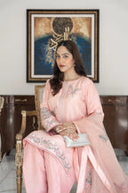 A woman seated in an ornate chair wearing a light pink traditional embroidered outfit with floral detailing, holding a small silver clutch in front of a framed artwork and dark wood cabinet.