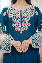 A close-up of a teal blue embroidered tunic top with ornate floral and paisley embroidery in beige, pink, and white, featuring a round neckline, decorative front buttons, and wide bell sleeves with scalloped cuffs. A person's hands are visible at the bottom of the frame.
