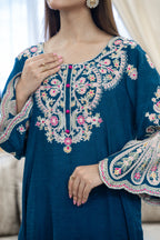 A close-up of a woman wearing a teal blue embroidered tunic with intricate floral embroidery in pink, white, and gold on the chest and sleeves.