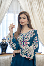 A woman wearing a teal blue embroidered kurta with intricate floral patterns in beige, pink, and green, standing in front of cream-colored curtains.