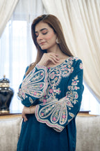 Woman wearing a blue embroidered dress indoors with white curtains.