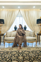 A woman wearing a brown embroidered traditional outfit sits on a cream-colored sofa in an elegant living room with gold-trimmed furniture, patterned curtains, and a large ornate rug.
