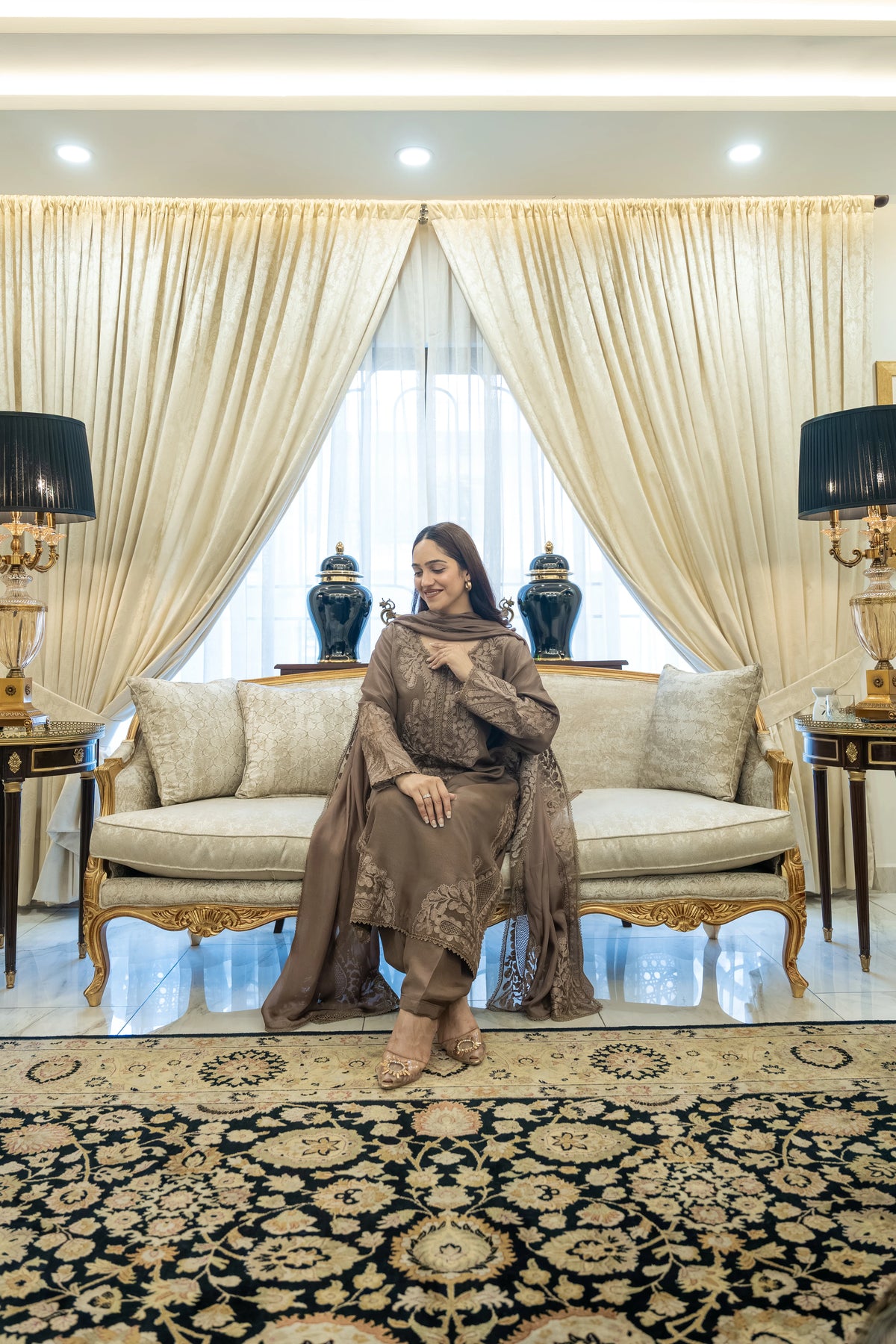A woman wearing a brown embroidered traditional outfit sits on a cream-colored sofa in an elegant living room with gold-trimmed furniture, patterned curtains, and a large ornate rug.