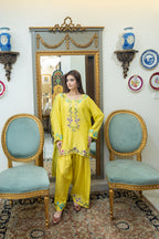 A woman modeling a bright yellow three-piece ethnic suit with colorful floral embroidery, standing in a decorated room with a large ornate mirror, two light blue chairs, and a patterned rug.