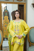A woman modeling a bright yellow traditional South Asian outfit with colorful floral embroidery on the neckline and sleeves, standing in front of an ornate gold-framed mirror in an indoor setting.