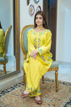 A woman sits on an ornate gold-framed chair wearing a bright yellow embroidered kurta set with matching pants and flat shoes, in an indoor room with a mirror, wall decor, and patterned rug.