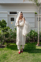 A woman wearing a traditional white embroidered kurta set with a matching sheer dupatta and white pants, standing on a green lawn in front of a white house.