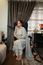 A woman sits in a dark blue armchair wearing a light blue traditional outfit with a matching dupatta. The setting is an indoor room with patterned curtains, a patterned rug, and dark wooden furniture.