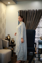 A woman wearing a light blue long kurta with floral lace embroidery on the hem and sleeves, standing in a furnished room with patterned wallpaper, curtains, and a sofa.