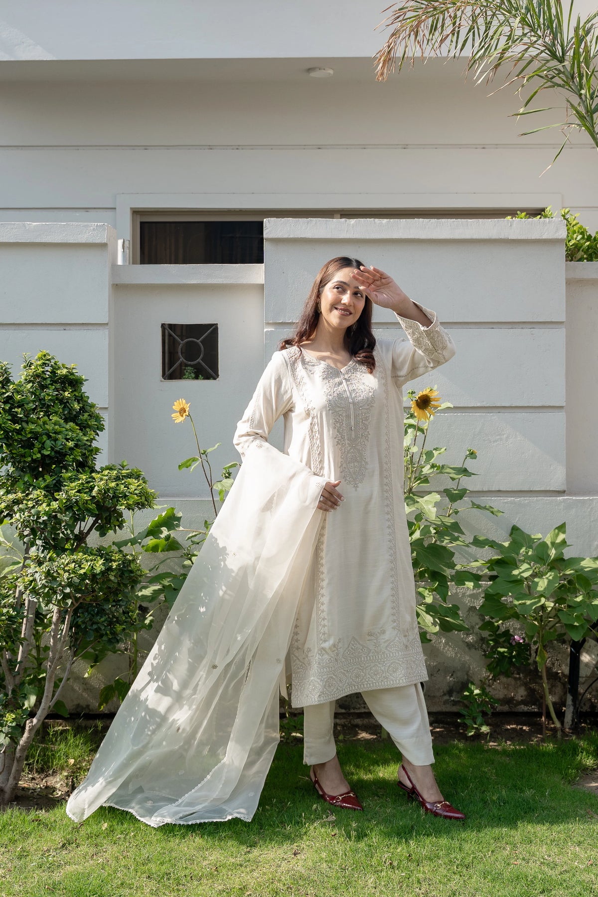 A woman modeling a traditional off-white embroidered kurta set with matching pants and dupatta, standing outdoors on a green lawn in front of a white house with sunflowers.