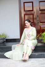 A woman sits on black stone steps in front of a dark wooden door, wearing a light sage green traditional outfit with pink floral embroidery and lace details.