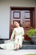 A woman sits on the steps in front of a dark brown wooden door, wearing a light sage green traditional outfit with pink floral embroidery.