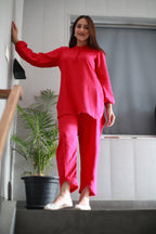A woman wearing a bright pink two-piece linen pajama set consisting of a long-sleeve top and matching pants, standing on a dark staircase in an indoor setting with a potted plant in the background.