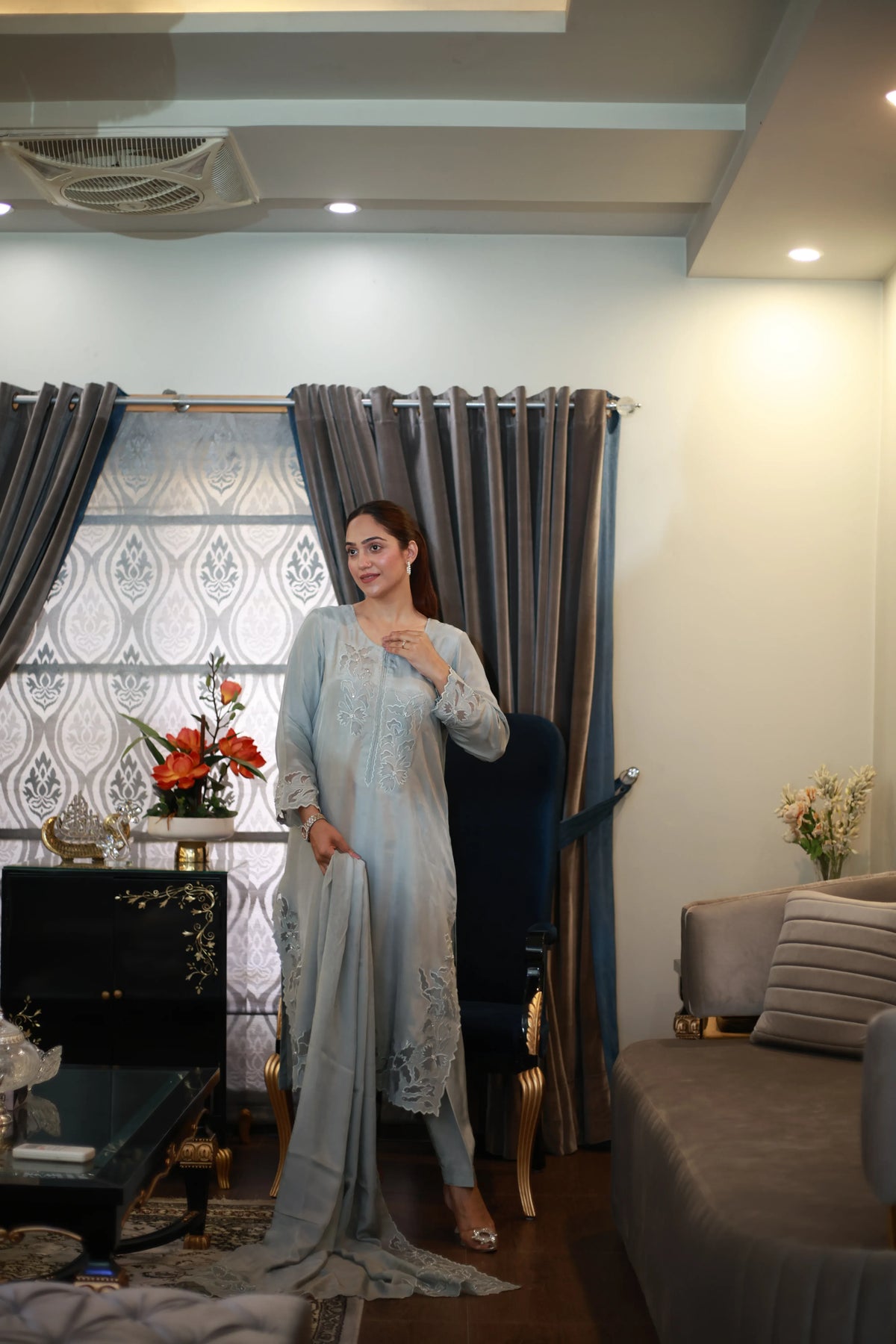 Woman wearing light grey embroidered 3-piece suit with dupatta, side view pose in living room