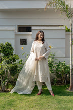 A woman wearing a traditional off-white embroidered kurta set with a matching dupatta and trousers, standing on a green lawn in front of a modern white house.