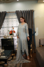 A woman modeling a light blue-grey traditional South Asian outfit with matching dupatta in an indoor living room setting with curtains and furniture.