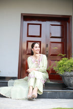 A woman wearing a light sage green traditional outfit with pink floral embroidery sits on the steps in front of a dark reddish-brown wooden door.